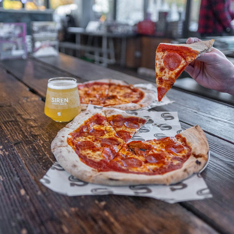 Person holding a slice of pizza with a beer on a wooden table in a casual setting.